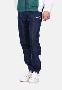Pantaloni da jogging blu scuro con una texture liscia, design affusolato, dettagli bianchi e un logo sulla gamba sinistra. Abbinati a sneaker bianche.