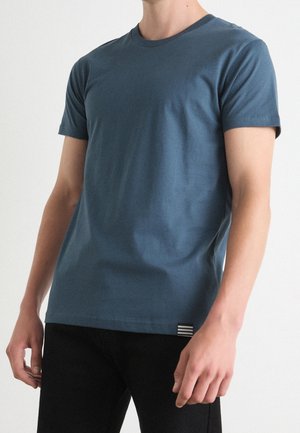Mężczyzna w jednolitym niebieskim t-shircie z krótkim rękawem i czarnych spodniach, stoi z ramionami swobodnie opuszczonymi wzdłuż ciała na białym tle.