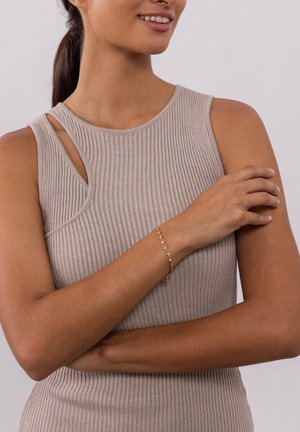 Beige ribbed tank top with a unique cutout design, paired with a gold chain bracelet featuring a small charm. Smooth textures evident.