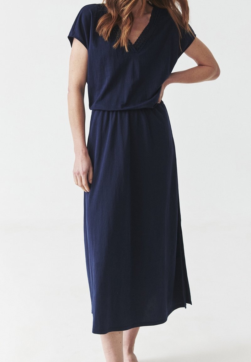 Femme portant une robe bleu marine à manches courtes avec un col en V et une jupe longueur cheville, debout avec une main sur la hanche sur fond blanc.