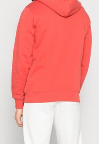 Personne portant un sweat à capuche corail et un pantalon blanc, vu de dos contre un fond clair.