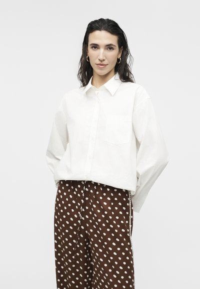 Femeie cu păr închis la culoare poartă o cămașă albă cu nasturi și pantaloni largi maro cu buline albe, stând într-un decor simplu, cu fundal deschis la culoare.