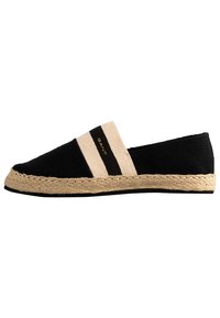 Espadrille en toile noire avec une semelle en jute tissée. Caractéristiques d'une bande élastique beige avec des rayures noires et un marquage doré sur le côté.