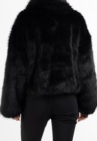 Schwarze Kunstpelz-Cropped-Jacke mit einer strukturierten, flauschigen Oberfläche und weiten Ärmeln. Kombiniert mit schwarzen, eng anliegenden Hosen.