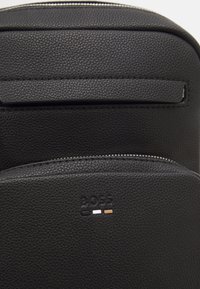 Sac à dos en cuir noir texturé avec un panneau supérieur lisse, fermeture à glissière, logo en relief et couleurs d'accent minimales en blanc et or.