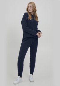 Granatowy sweter w prążki oraz pasujące spodnie w luźnym kroju. Sweter ma długie rękawy i opadające ramiona. Całość dopełniają białe trampki.