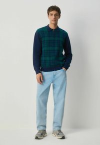 Grüner und navyblauer, karierter Strickpullover mit Kragen, kombiniert mit hellblauen, geraden Jeans und grauen Sneakers.