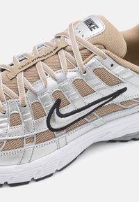 Chaussure de course Nike beige et argentée avec tissu mesh, superpositions en couches, logo Swoosh noir, semelle blanche et texte « Boytearn Series » sur les lacets.