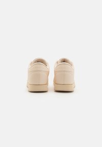 Beige faux leather sneakers with a smooth texture, low profile, rounded heel, and embossed branding on the back. Simple design, minimal accents.