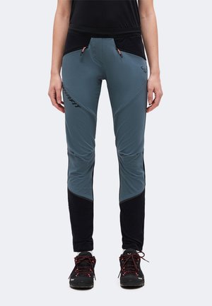 Persona che indossa pantaloni da esterno bicolore blu e nero con tasche con zip e scarpe da trekking nere con lacci rossi, in piedi.