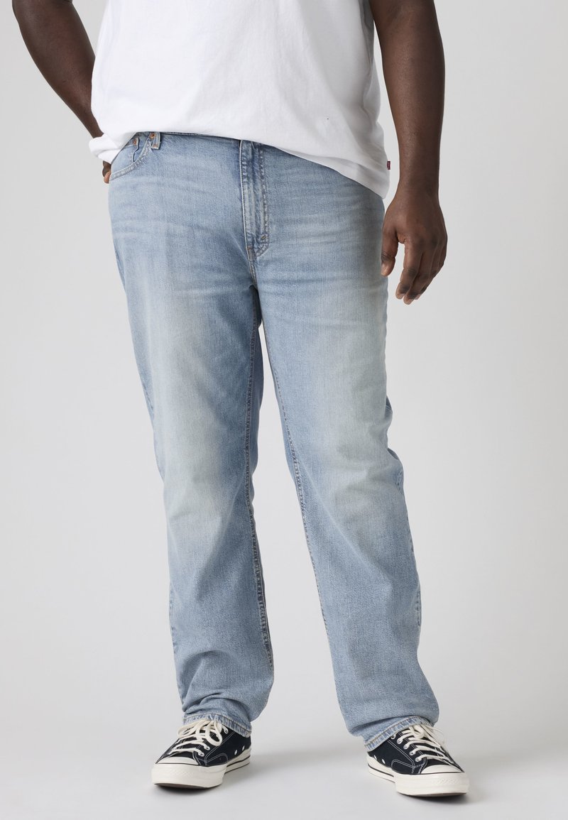 Άτομο που φοράει ελαφρώς μπλε τζιν, μαύρα και λευκά αθλητικά παπούτσια και λευκό μπλουζάκι, στέκεται με το ένα χέρι στην τσέπη.