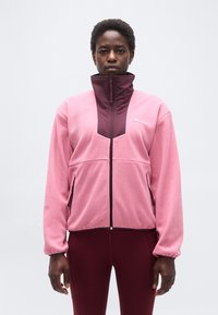 Ενήλικας που φοράει ροζ και μπορντό φλις ζακέτα με φερμουάρ και ψηλό γιακά, μαζί με μπορντό κολάν, στέκεται μπροστά σε απλό ανοιχτό φόντο.