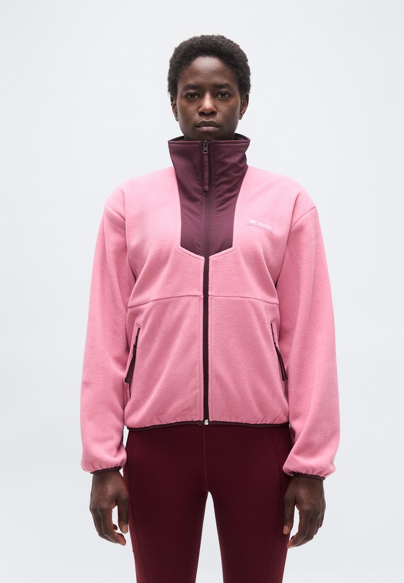 Ενήλικας που φοράει ροζ και μπορντό φλις ζακέτα με φερμουάρ και ψηλό γιακά, μαζί με μπορντό κολάν, στέκεται μπροστά σε απλό ανοιχτό φόντο.