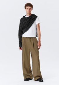 Schwarzer Strickpullover, der über einem weißen T-Shirt hängt, kombiniert mit lockeren, beigen Hosen und schwarzen Schuhen, mit minimalistischem Design und lässiger Passform.