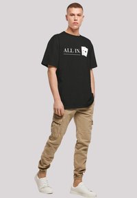Schwarzes Baumwoll-T-Shirt mit "ALL IN."-Text und Spielkarten-Print auf der Brust, kombiniert mit khakifarbenen Cargo-Hosen und weißen Sneakers.