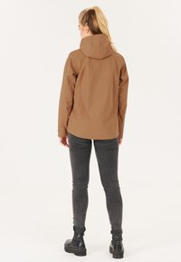 Brun vattentät jacka med huva, med en slät textur och en något längre bak. Matcherade med grå åtsittande jeans och svarta stövlar.