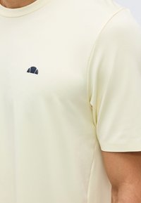 Άνδρας που φοράει κρεμ κοντομάνικη μπλούζα με μικρό μαύρο ημικύκλιο λογότυπο στην αριστερή πλευρά του στήθους.