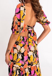 Robe maxi avec des manches bouffantes et un dos nu, présentant un motif floral en noir, rose, orange et jaune. Liens à la taille.