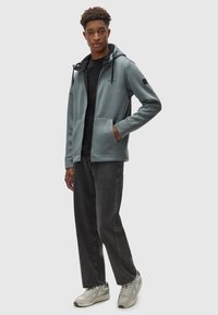 Hellblauer Reißverschluss-Hoodie mit strukturiertem Oberflächen, Fronttaschen und Bindebändern; getragen mit schwarzem Shirt, dunkler Jeans und grauen Sneakers.