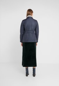 Woman wearing a navy quilted jacket, gray turtleneck, long dark green skirt, and navy heeled ankle boots, standing facing away on plain background.