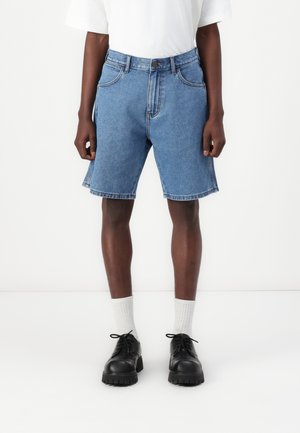 Denim shorts vaaleansinisinä, suoran leikkauksen, viiden taskun muotoilun ja etunapilla. Yhdistetty mustiin kenkiin ja valkoisiin sukkiin.