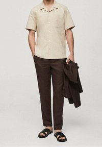 Beige overhemd met korte mouwen en een borstzak, bruine slim-fit broek en zwarte sandalen. Licht materiaal en ontspannen ontwerp voor warm weer.