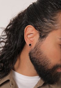 Cercel negru circular, din metal, purtat în lobul urechii. Părul ondulat negru și barba scurtă sunt parțial vizibile pe un fundal neutru.