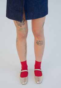 Sidabriniai Mary Jane bateliai su dirželiu, derinami su raudonomis raištinėmis kojinėmis. Atskleistos kojos pademonstruoja įvairius juodus ir žalius tatuiruotes. Matomas mėlynų džinsinių sijonų.