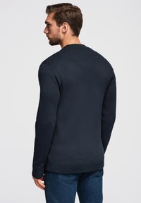 Dunkelblauer Rundhalsausschnitt-Pullover aus strukturiertem Stoff mit gerippten Ärmeln und Saum. Die Rückseite hat eine dezente Nahtdetails. Über blauen Jeans getragen.