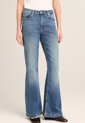 Blaue hochtaillierte Schlagjeans mit Fronttaschen und Knopfverschluss, getragen mit schwarzen Ballerinas vor neutralem Hintergrund.