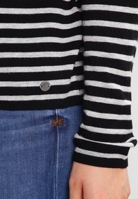 Pull en maille rayé noir et blanc avec un ourlet arrondi, orné d'un détail logo argenté près du bas et de poignets côtelés. Porté sur un jean.