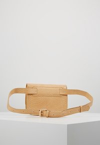 Béžová ledvinka s imitací hadí kůže, nastavitelným popruhem a zlatou sponou, vystavená na bílé ploše před jednoduchým šedým pozadím.