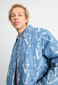 Persona con cabello rizado rubio que lleva una chaqueta de mezclilla azul con un patrón de alambre de espino y diseños de la letra V sobre un fondo claro.