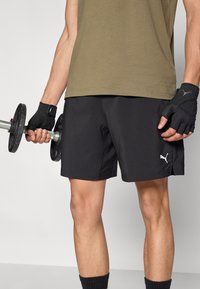 Μαύρα αθλητικά σορτς με ελαφρύ σχεδιασμό και ασημένιο λογότυπο Puma. Το μοντέλο κρατά ένα βαράκι, φοράει γάντια χωρίς δάχτυλα και πράσινο πουκάμισο.