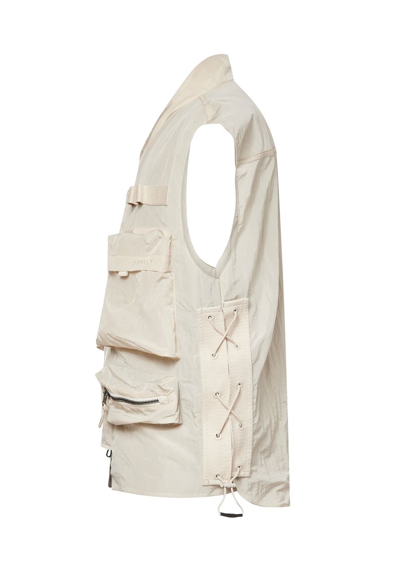 Chaleco utilitario sin mangas de color beige con gran bolsillo delantero con solapa, bolsillo inferior con cremallera, cordones laterales ajustables y amplias sisas.
