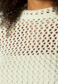 Stickad tröja med ett öppet, diamantmönstrat väv i krämfärg, med en texturerad yta och en solid, ribbad halsringning.