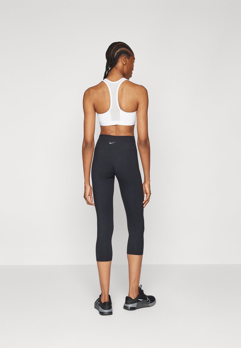 Sostén deportivo blanco de tirantes en la espalda y leggings capri negros con cintura alta. Zapatillas deportivas negras con acentos grises. Fondo plano.