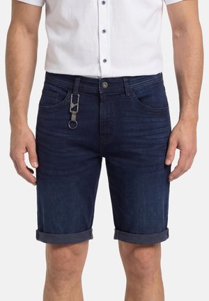 Mann trägt dunkelblaue, hochgekrempelte Jeansshorts mit Knopf- und Reißverschluss, Vordertaschen, und ein weißes, kurzärmeliges Hemd mit Knöpfen.