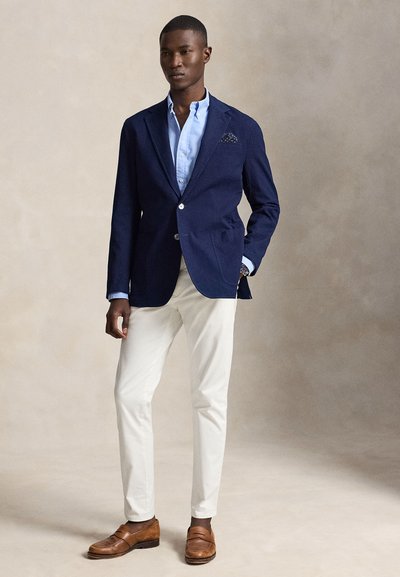 Homme portant un blazer bleu marine, une chemise bleu clair, un pantalon blanc et des mocassins marron, debout avec une main dans la poche.