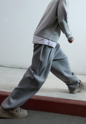 Pull gris avec des poignets côtelés, superposé sur une chemise blanche, associé à un pantalon de survêtement gris ample avec des plis. Baskets beige clair.