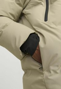 Beige puffer jacket featuring a textured exterior, black cuff, and a zippered pocket on the chest. Soft, insulated fabric for warmth.
