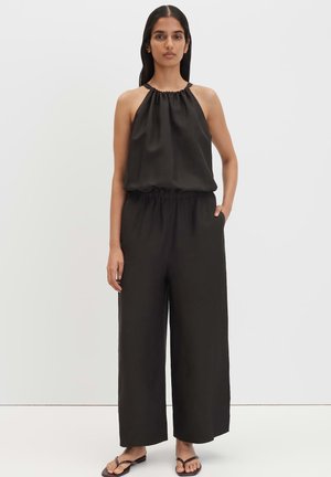 Frau steht und trägt einen ärmellosen schwarzen Halter-Jumpsuit mit weitem Bein und flachen Sandalen vor schlichtem weißem Hintergrund.