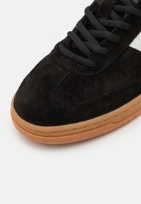 Adidași negri din suede cu o textură netedă, având șireturi negre și o talpă din cauciuc caramel. Accente fine de cusătură de-a lungul marginii.