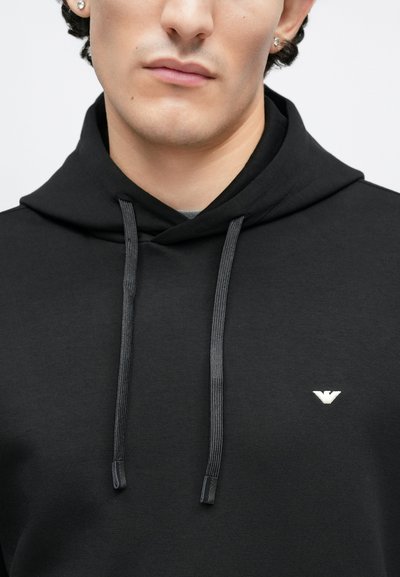Melnas kapuces džemperis no gludas auduma, ar nelielu baltu logotipu uz krūškurvja un kontrastējošām pelēkām auklām.