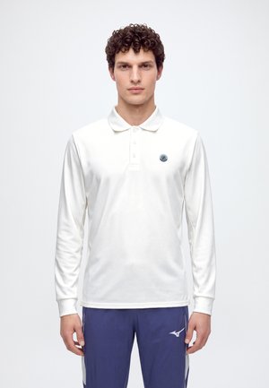 Un homme porte un polo blanc à manches longues avec un emblème sur la poitrine et un pantalon de sport bleu, sur un fond uni et clair.