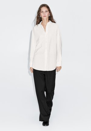 Camicia bianca a righe con un taglio rilassato, abbinata a pantaloni neri larghi, presentata su uno sfondo neutro.