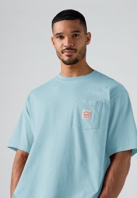 Άνδρας με κοντά μαλλιά και γενειάδα, φορώντας ένα ανοιχτό μπλε φαρδύ T-shirt με ένα μικρό κόκκινο και λευκό έμβλημα στην τσέπη του στήθους.