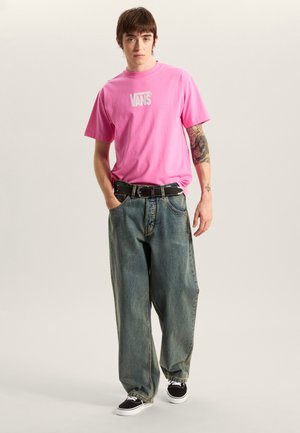 Camiseta de algodón rosa con el logo "VANS" en blanco, combinada con unos vaqueros azules descoloridos y holgados, y zapatillas negras con suelas blancas.