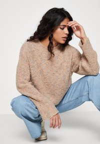 Maglione lavorato a maglia in morbido beige con macchie multicolori, scollo rotondo e spalle scese. Abbinato a jeans azzurri chiari.