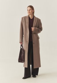 Langer, beiger Wollmantel mit Reverskragen, Ein-Knopf-Verschluss und Seitentaschen. Trägt darunter ein dunkles Hemd und schwarze Hose.